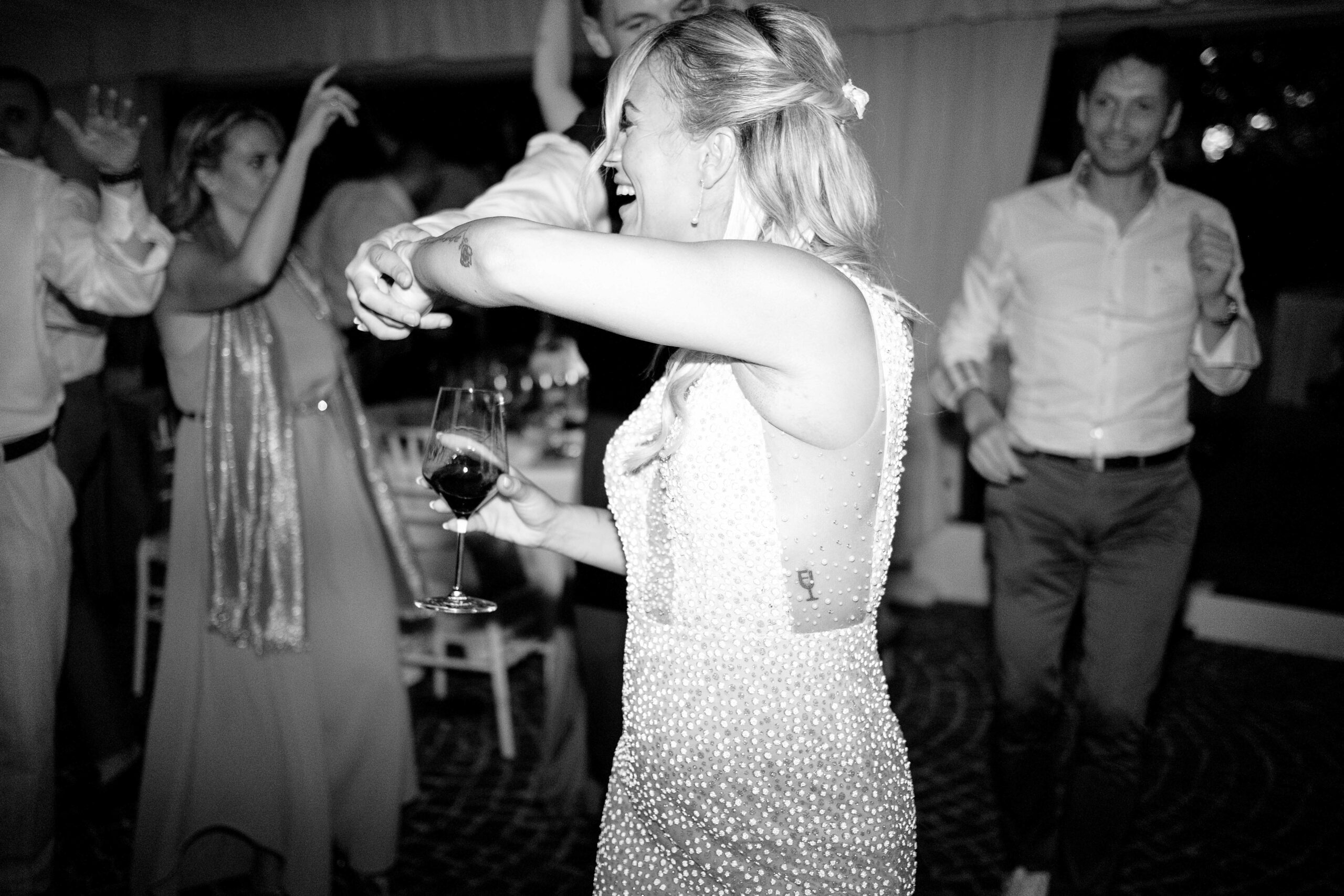 Una donna con un abito di perline sorride e balla in un vivace evento al chiuso, tenendo in mano un bicchiere di vino. Altre persone ballano e ridono sullo sfondo. La scena è gioiosa ed energica.