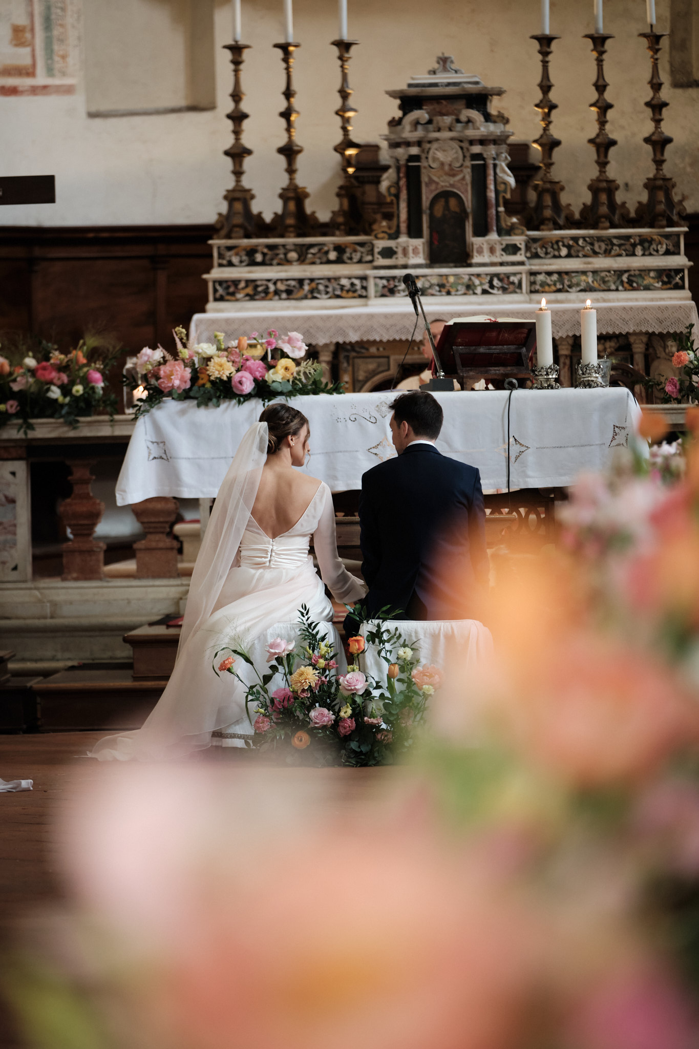 Una sposa e uno sposo si inginocchiano fianco a fianco sull'altare di una chiesa decorata, circondati da composizioni floreali e candele, durante la loro cerimonia nuziale.