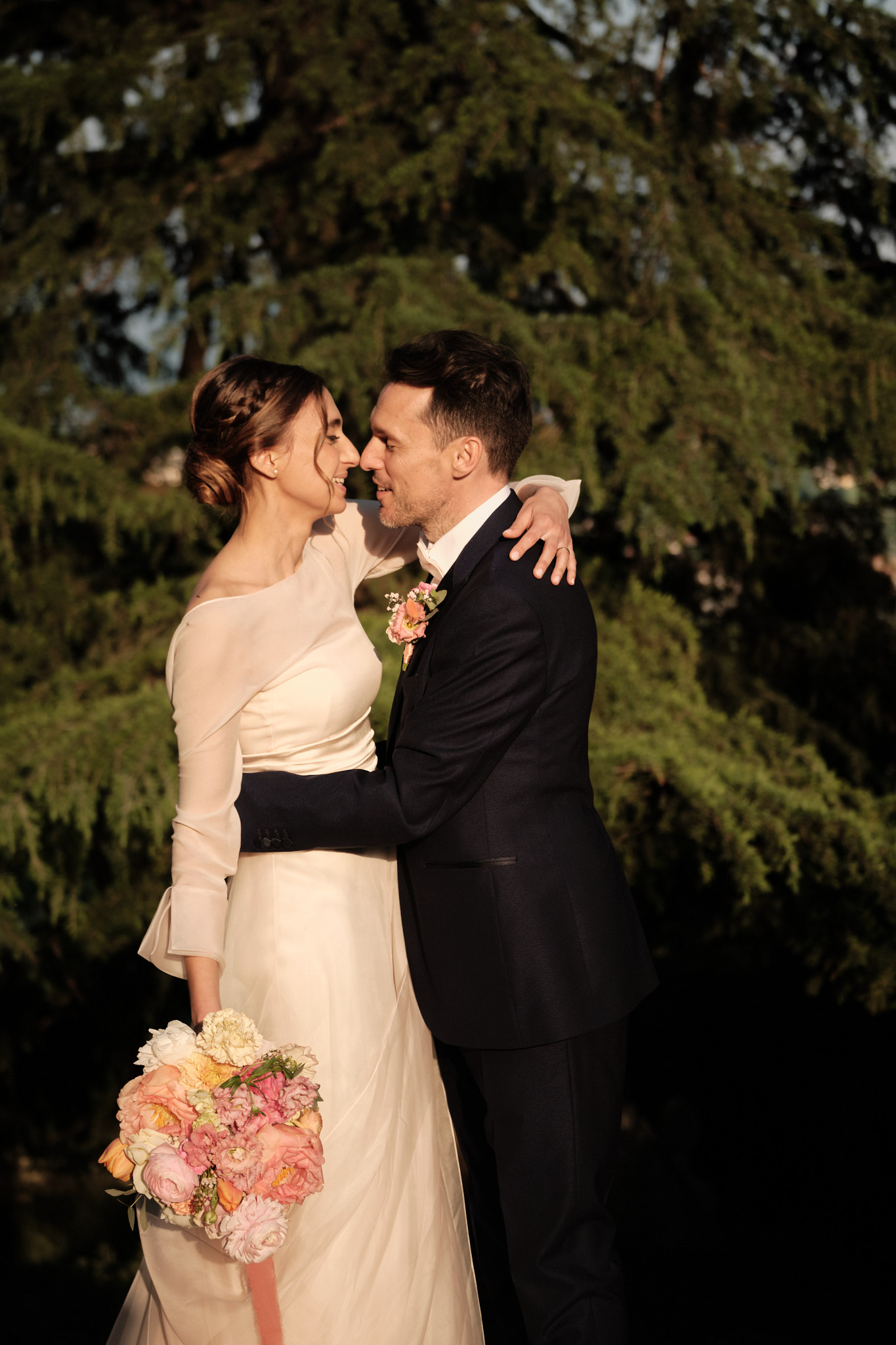 Una sposa e uno sposo all'aperto, sorridenti e abbracciati. La sposa tiene in mano un bouquet di fiori pastello ed entrambi sono vestiti in abiti da cerimonia con alberi sullo sfondo.