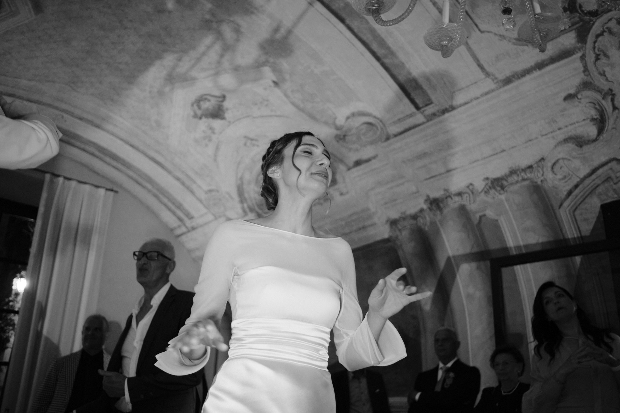 Una donna in abito bianco sorride e gesticola gioiosamente sotto un soffitto a volta ornato, circondata da persone in abiti formali in quello che sembra essere un evento celebrativo. L'immagine è in bianco e nero.