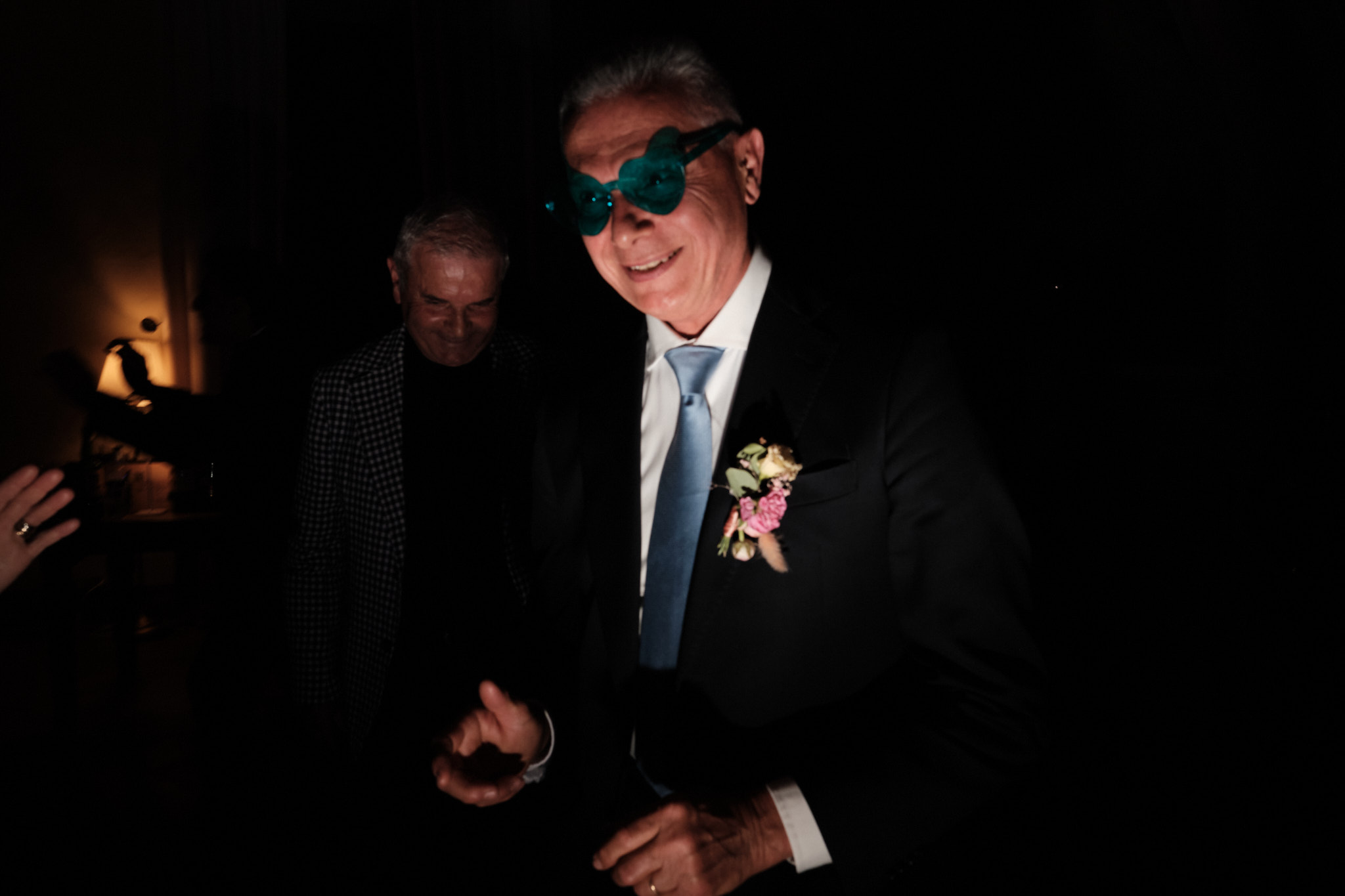 Un uomo anziano in abito e cravatta blu, con occhiali blu oversize e boutonniere, sorride in una stanza poco illuminata con un altro uomo sullo sfondo.