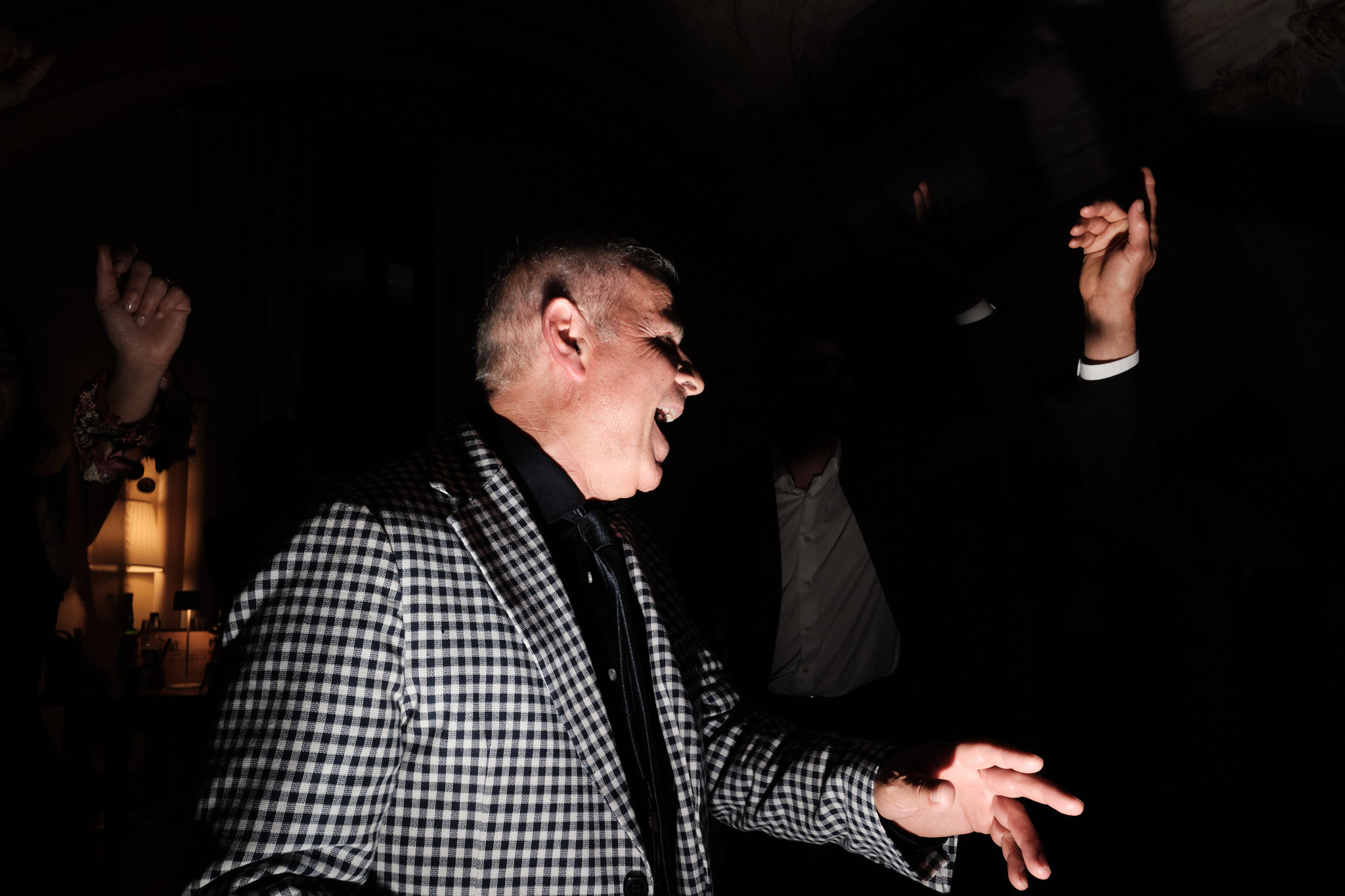 Un uomo con una giacca a scacchi sorride e gesticola con le mani mentre balla in una stanza poco illuminata, circondato da altre persone le cui mani sono visibili nell'ombra.