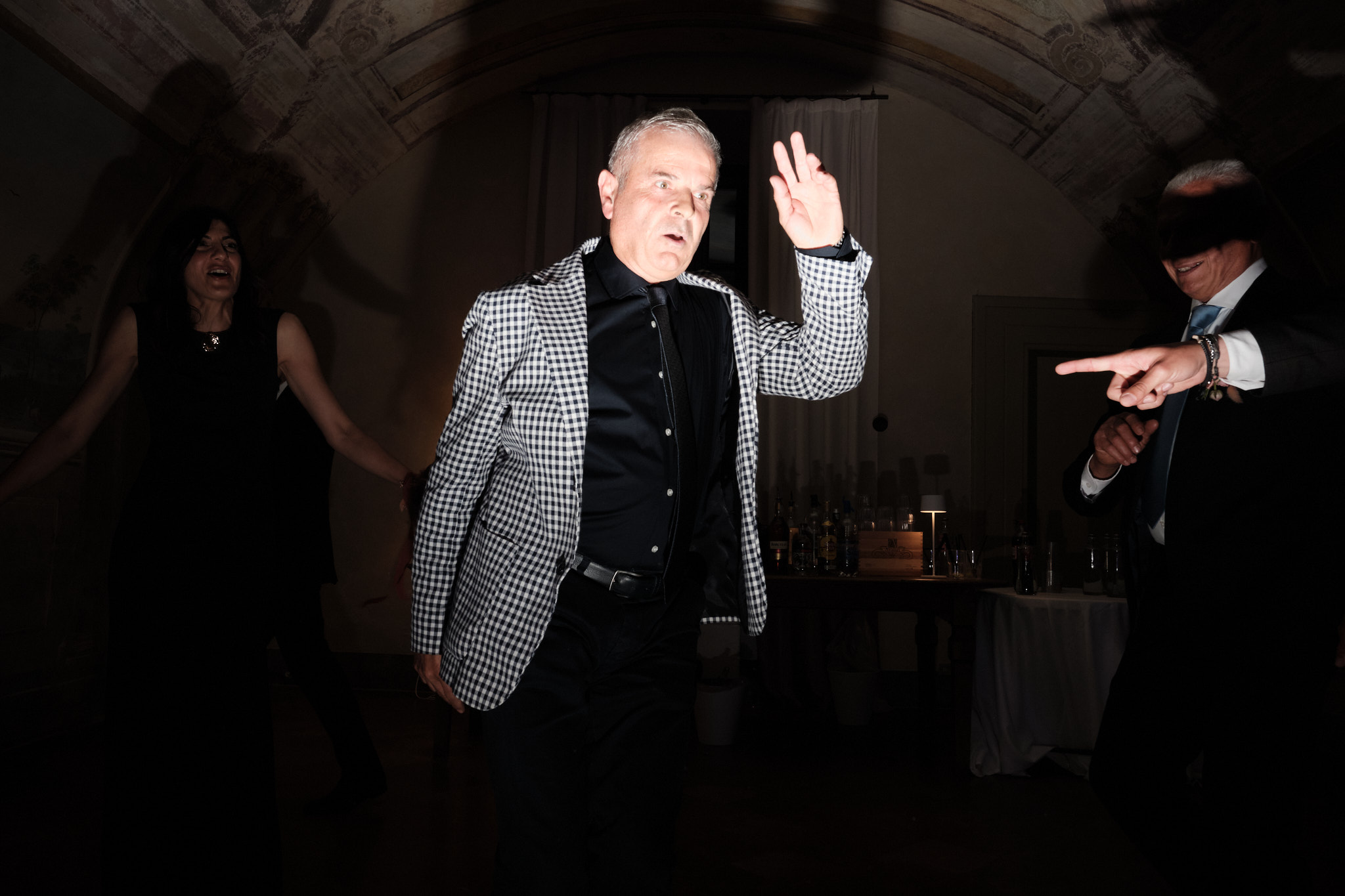 Un uomo con una giacca a scacchi balla energicamente sotto un'illuminazione drammatica in un evento al chiuso, con altre due persone in abiti scuri che ballano sullo sfondo.