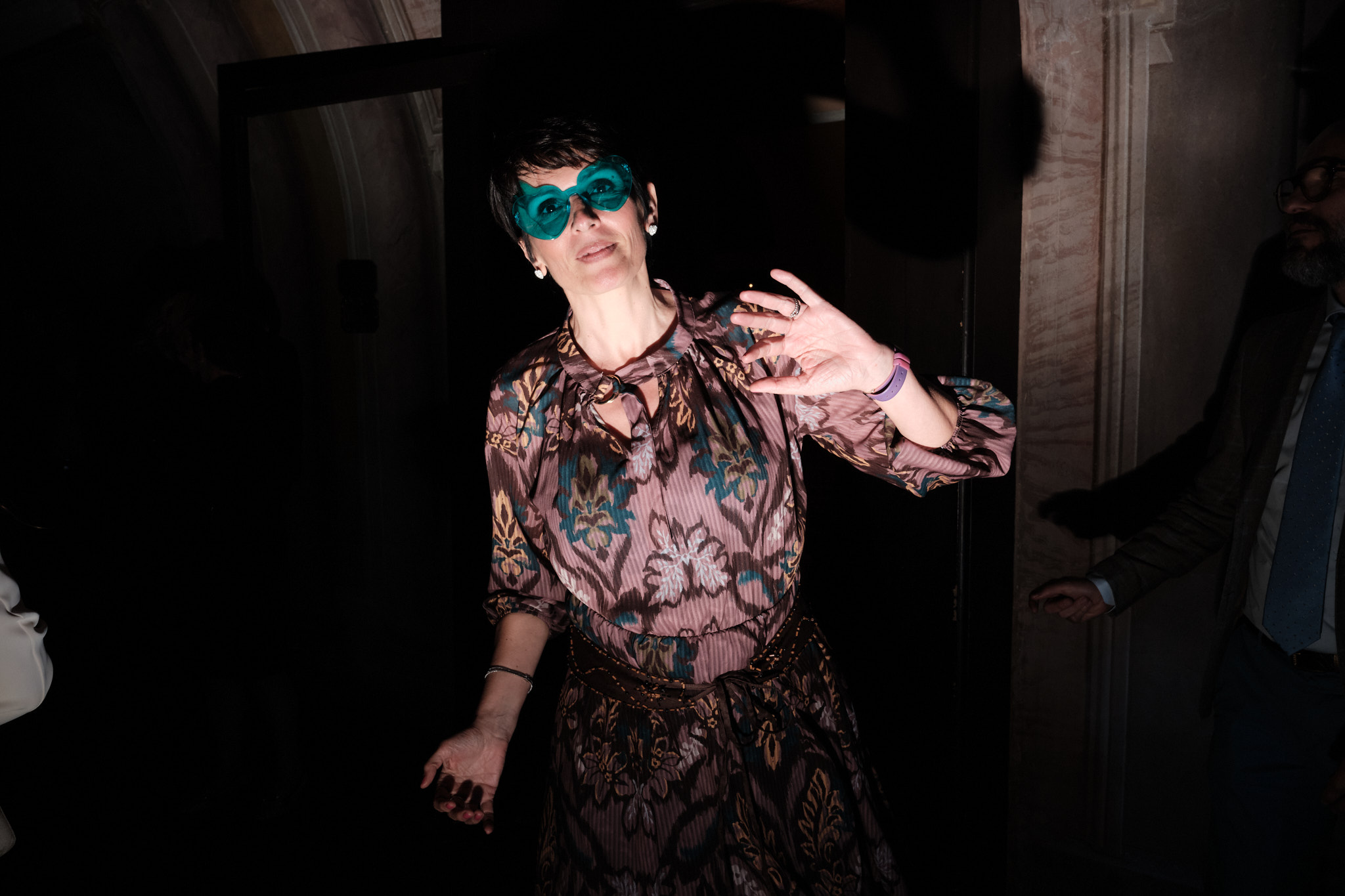 Una donna con un abito a fantasia e una maschera verde acqua gesticola con le mani in un'illuminazione drammatica, in piedi in un interno con uno sfondo in ombra.