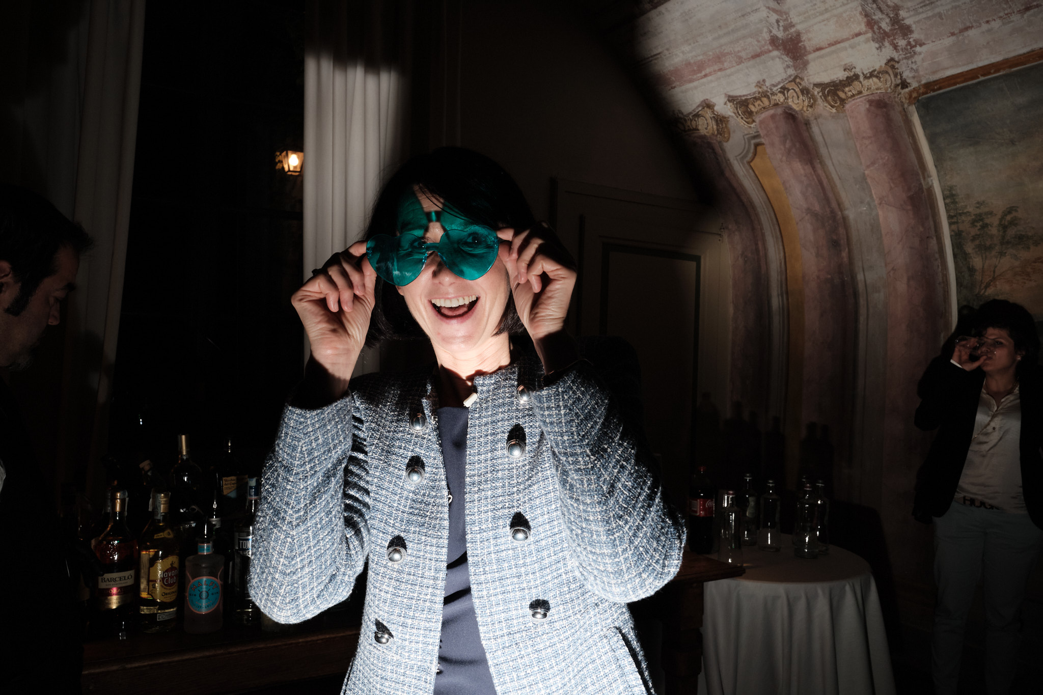 Una donna con una giacca a fantasia sorride e regge degli occhiali azzurri in una stanza poco illuminata, con delle bottiglie su un tavolo alle sue spalle e un'altra persona che beve sullo sfondo.