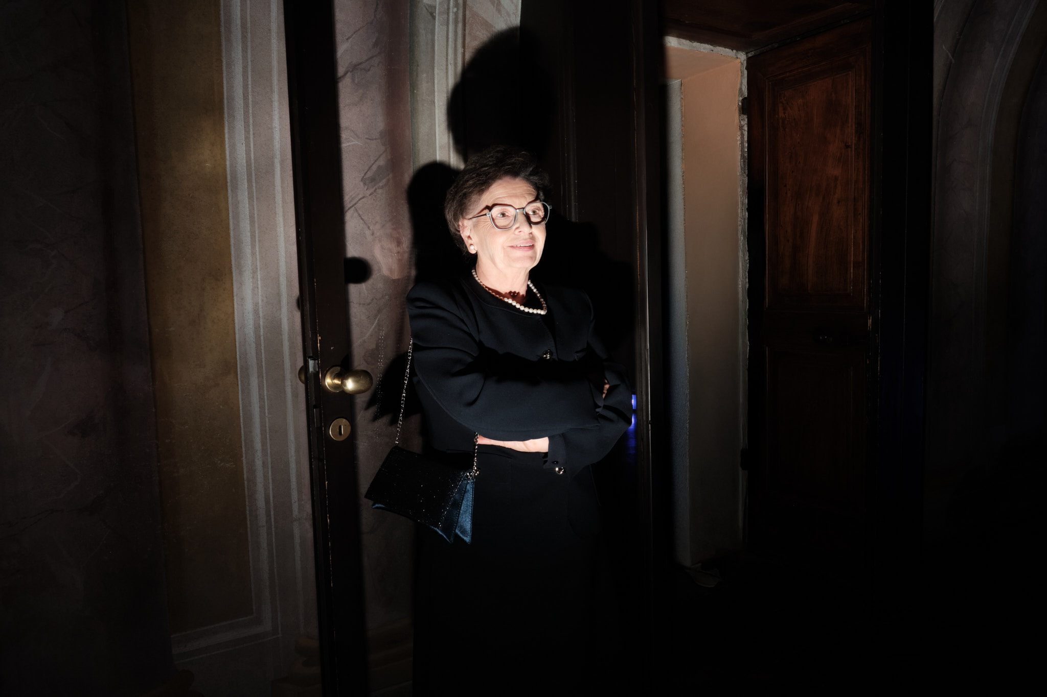 Una donna anziana che indossa occhiali, un abito nero e una collana di perle sta in piedi con le braccia incrociate in una stanza poco illuminata, proiettando un'ombra sulla parete alle sue spalle.